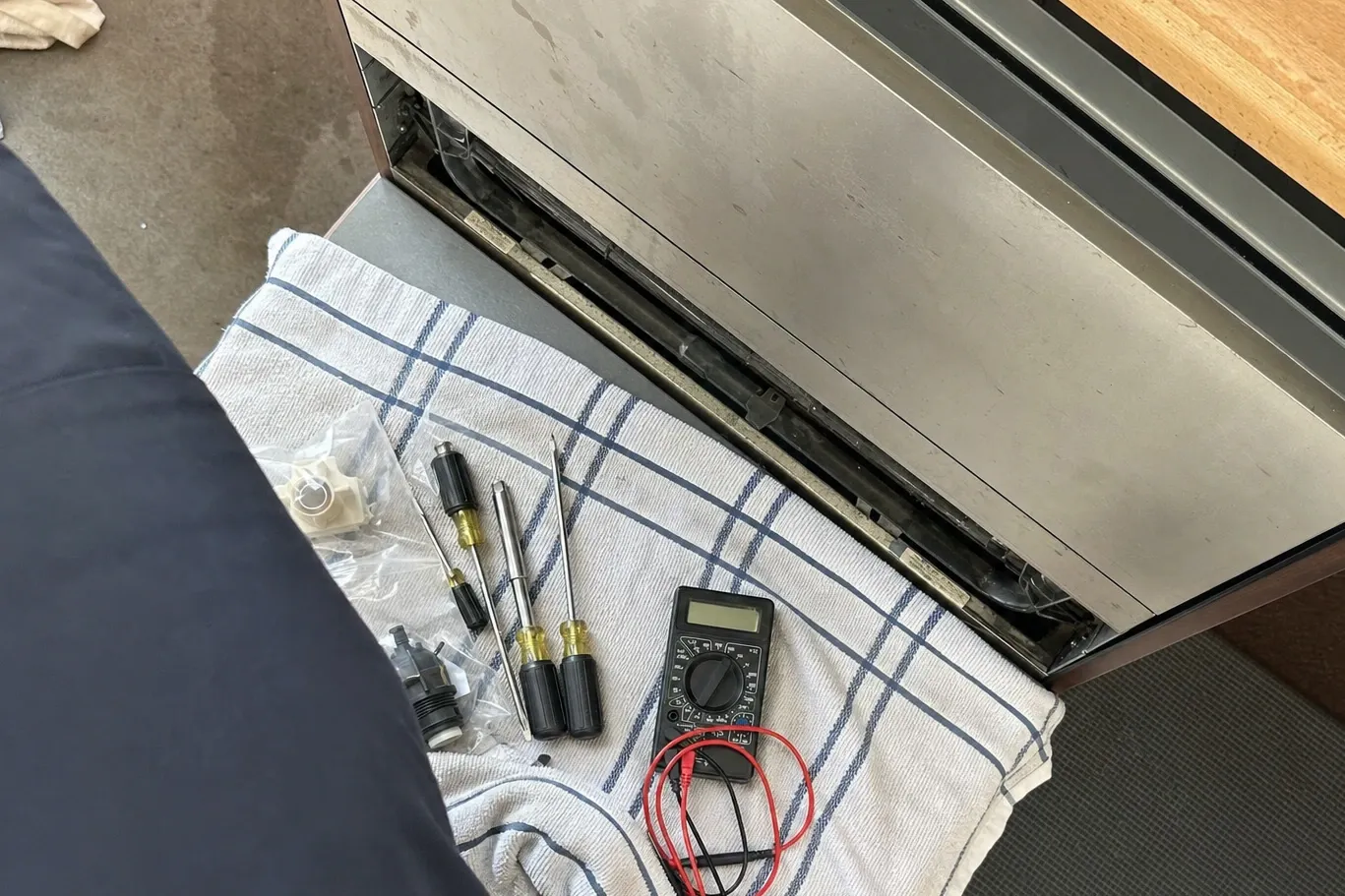 Two bagged replacement valves, four screwdrivers and a multimeter with coiled leads laid out on a towel beside the opened kick panel of a stainless built-in