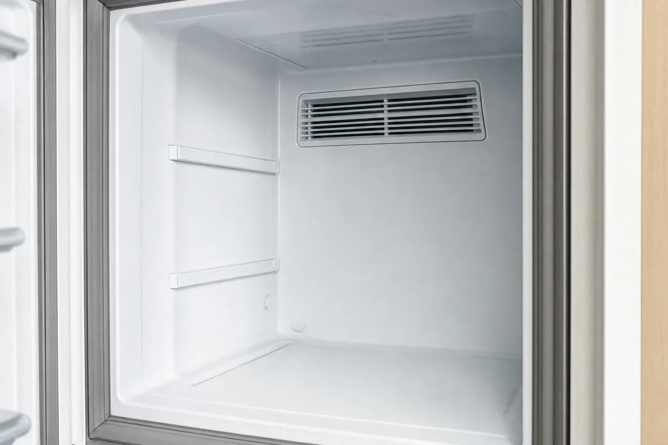 The empty white interior of a built-in refrigeration compartment, with two shelf supports and a louvered air-duct grille high on the rear wall